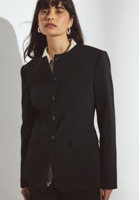 Juodas pritaikytas švarkas su aukštu apykakle, mygtukų uždarymu ir šoninėmis kišenėmis. Skaidri tekstūra, tvirtas siluetas ir paprasti dizaino akcentai.