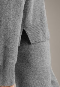 Grau wöllener Pullover mit geripptem Bund, der ein einzigartiges seitliches Schlitz-Detail aufweist. Die Textur ist weich und das Material wirkt warm und gemütlich.