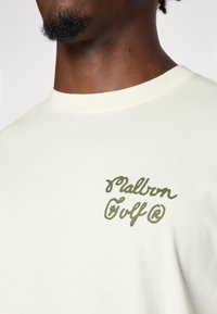 Cremefarvet bomulds t-shirt med et grønt broderet logo på venstre bryst, der står "Malbon Golf." Klassisk rund halslinning og blød tekstur.