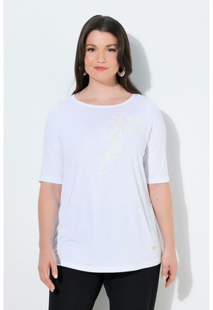 Frau mit langen dunklen Haaren, die ein weißes Kurzarmshirt mit dezentem Blumendesign, schwarze Hose und Creolen trägt, vor weißem Hintergrund stehend.