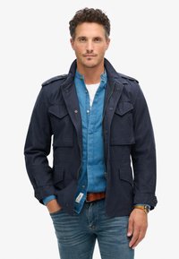 Mørkeblå bomuldsjakke med to brystlommer, skulderklapper og lynlås, lagd over en lyseblå skjorte med knapper, parret med jeans.