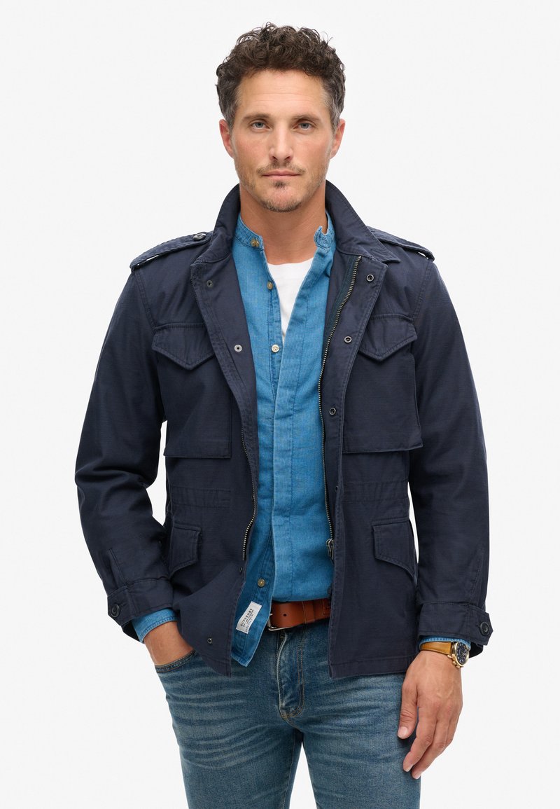 Mørkeblå bomuldsjakke med to brystlommer, skulderklapper og lynlås, lagd over en lyseblå skjorte med knapper, parret med jeans.