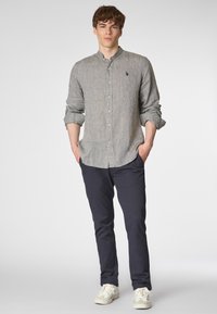 Giovane uomo con capelli ricci è in piedi con sicurezza, indossando una camicia grigia chiara con bottoni e pantaloni blu scuro, con le mani nelle tasche, su uno sfondo neutro.