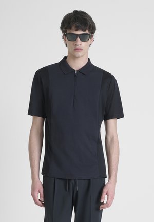 Zwarte polo met korte mouwen, voorrits, gemêleerde stof en zijpanelen. Gecombineerd met zwarte broek en een donkere zonnebril.