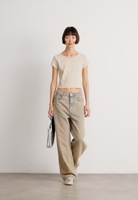 Beige avkortet T-skjorte med korte ermer, parret med lyse beige vide jeans. Modellen holder en svart veske og har på seg sølvfargede sneakers.