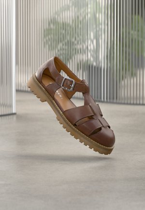 Sandale maro din piele cu vârf închis, cu multiple curele, cataramă argintie și talpă texturată din cauciuc, prezentată plutind deasupra unui podea de beton.