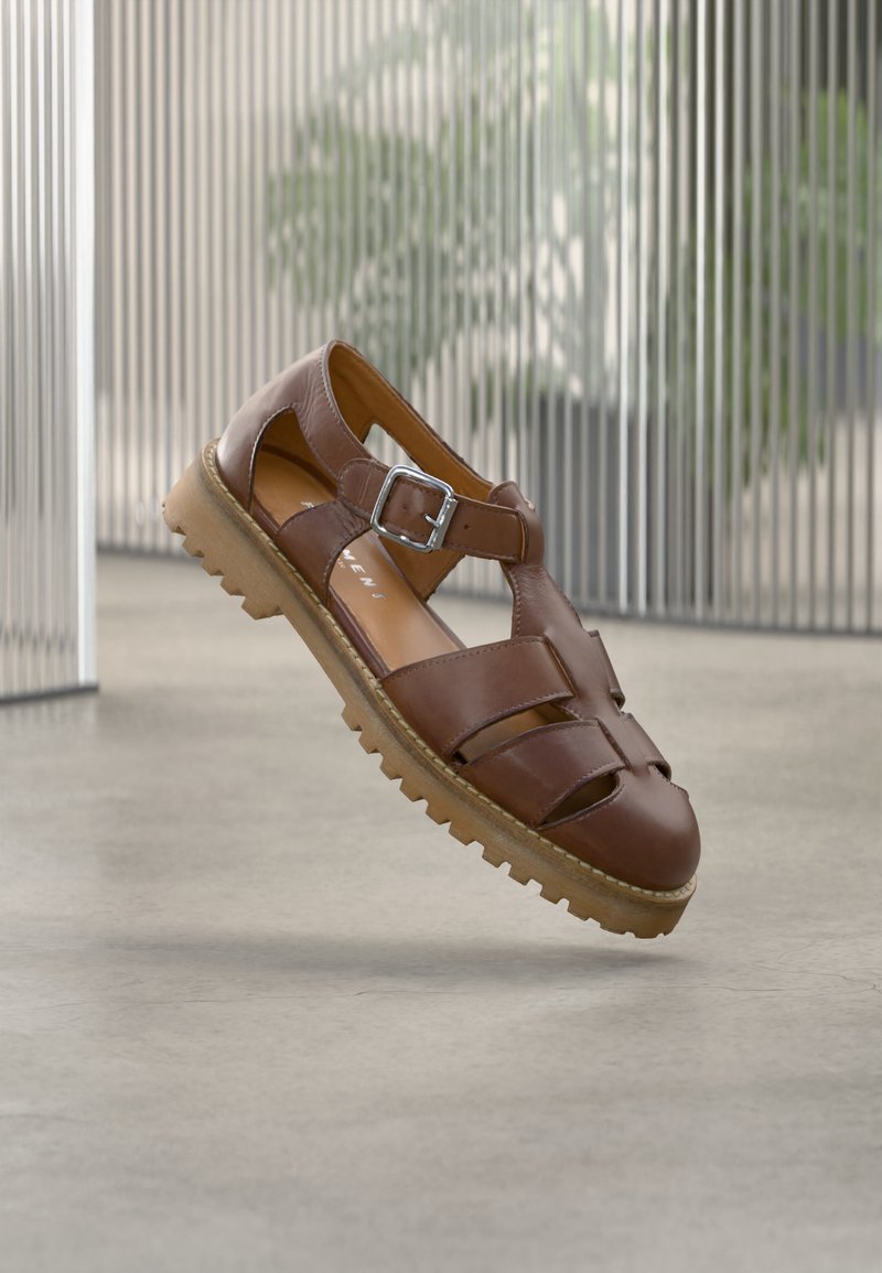 Pruun nahast kinnise varbaga sandaal mitme rihmaga, hõbedase pandla ja tekstuurse kummist tallaga, mis on kujutatud betoonpõranda kohal hõljumas.