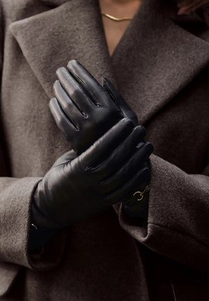 Zwarte leren handschoenen met een gladde textuur, voorzien van een gestikt ontwerp en een kleine metalen accenten, tegen een bruine jas gehouden.