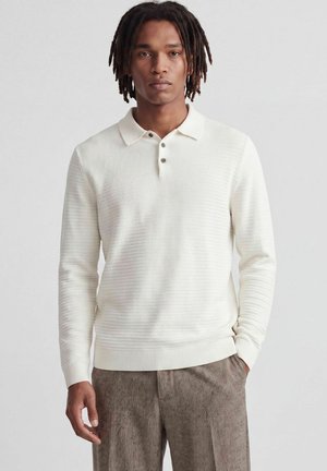 Jeune homme aux dreadlocks foncées portant un polo en maille crème à manches longues et un pantalon marron texturé, debout devant un fond uni.
