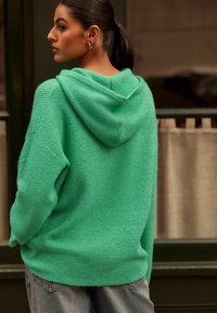 Roheline kudumine kapuutsiga, millel on reguleeritav pael, soonikkoes ja üleliigne lõige, koos helesiniste jeanidega.