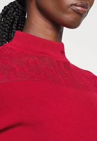 Maglione rosso lavorato a maglia con colletto alto. Presenta dettagli testurizzati sulle spalle e una silhouette aderente. Il materiale sembra morbido ed elastico.