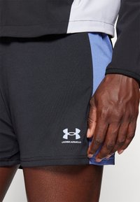 Melnas sporta bikses ar zilu sietu paneli, uz kuras redzama balta Under Armour logo. Audums ir elastīgs ar gludu tekstūru.