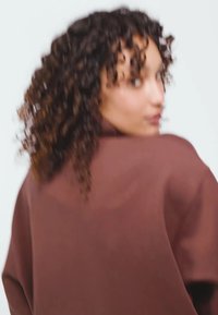 Hnedý oversized sveter s vysokým golierom, jemnou textúrou a širokými rukávmi, ktorý nosí modelka s kučeravými vlasmi, otočená mierne nabok.