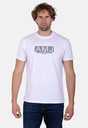 Hombre con el pelo rizado, que viste una camiseta blanca de AMJ Amaro Jeans y vaqueros azules, de pie de frente sobre un fondo claro y liso.