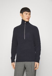 Marineblauer gerippter Pullover mit hohem Kragen und silbernem Reißverschluss. Strukturierter Stoff und taillierte Passform ergänzen die graue Hose. Neutraler Hintergrund.