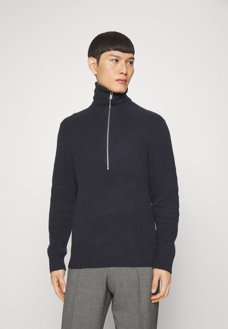 Marineblauer gerippter Pullover mit hohem Kragen und silbernem Reißverschluss. Strukturierter Stoff und taillierte Passform ergänzen die graue Hose. Neutraler Hintergrund.