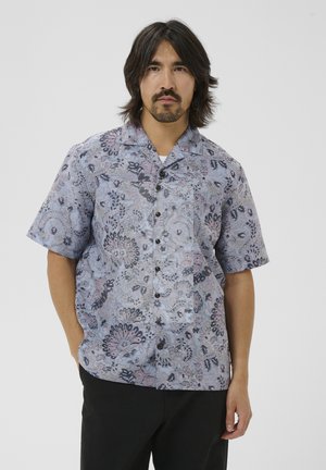 Mand med mellem-langt hår og overskæg, iført en kortærmet, knappet blå skjorte med et blomster- og abstrakt mønster.