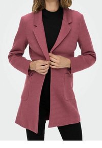 Femme portant un long blazer texturé rose poudré sur un haut noir et un pantalon noir, en train de boutonner les boutons avant du blazer.