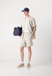 Conjunto de camisa de manga corta a rayas y pantalones cortos en crema y beige. El modelo lleva una gorra navy y sostiene un bolso navy, con zapatillas beige.
