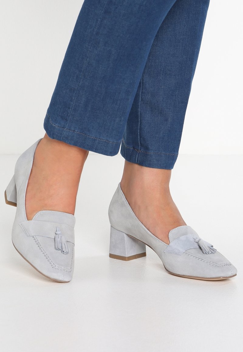 Mocassins à talons blocs en daim gris avec un bout pointu, présentant un gland décoratif. Les chaussures ont une texture lisse et un minimum de détails de couture.