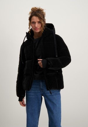 Zwarte pufferjack met een capuchon, ritsluiting en zijzakken. Beschikt over elastische mouwen en een matte afwerking. G gedragen over een donker top en blauwe jeans.
