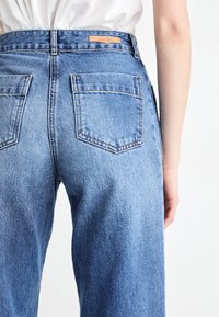 Jeans de mezclilla azul claro con cintura alta, dos bolsillos traseros con solapas puntiagudas y una sutil etiqueta de marca de cuero en la cintura.