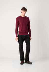 Bordeaux genser med lange ermer, rund halslinje og en liten logo detalj. Kombinert med svarte jeans og mørke sko.