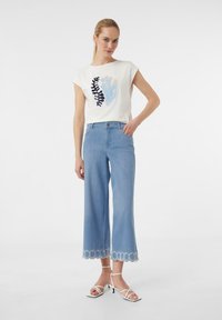 Weißes Kurzarmhemd mit floralem Stickerei, kombiniert mit hellblauen weit geschnittenen Jeans mit einem gewellten Spitzenabschluss und weißen Riemchensandalen.