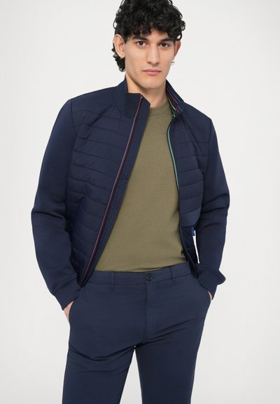 Tumesinine tepitud jope püstiäärisega ja mitmevärviliste tõmblukuga, mida on kantud vormitülikaga oliivrohelise särgi ja tumesiniste pükstega.