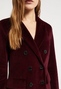 Bordová zamatová dvojradová sako s čiernymi gombíkmi, s klopami a bočnými vreckami. Textúrovaná tkanina s jemným leskom.