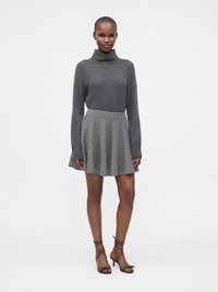 Femme debout portant un pull à col roulé gris foncé, une jupe plissée gris clair et des sandales à talons noires à brides sur fond blanc.