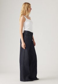 Φαρδιά τζιν παντελόνια σε σκούρο μπλε χρώμα, με ψηλή μέση, εμπρός τσέπες και ετικέτα στο πίσω μέρος. Κατασκευασμένα από ανθεκτικό τζιν ύφασμα.