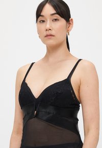 Musta alusvaatesetti, jossa on pitsireunus, läpinäkyvää kangasta, säädettävät olkaimet ja keinonahkainen vyötärönauha, jonka keskellä on leikattu yksityiskohta.