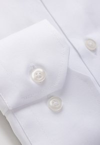 Valkoinen kauluspaita, jossa on sileä tekstuuri, terävä kaulus, hihansuut ja useita yksinkertaisia valkoisia nappeja. Saumat ovat näkyvissä reunoilla.