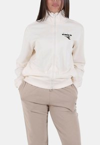 Giacca bianca con zip in tessuto morbido, presenta dettaglio logo nero, con polsini e fascia in vita a coste. Abbinata a pantaloni beige chiaro.