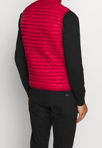 Červená vycpaná vesta s prošívaným designem a vysokým límcem, nošena přes černé tričko s dlouhým rukávem, v kombinaci s černými kalhotami a viditelným detailem zadní kapsy.