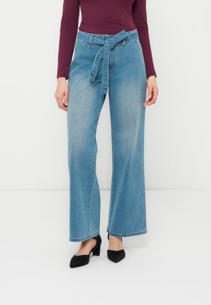 Ženska, oblečena v svetlo modre široke kavbojke s pasom, vezanim v vozel, bordo majico z dolgimi rokavi in črne koničaste pete, na belem ozadju.