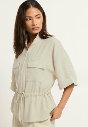 Kobieta w beżowej kurtce z krótkimi rękawami, z przednimi kieszeniami i paskiem w talii, patrzy w lewo na jednolity jasny tle.