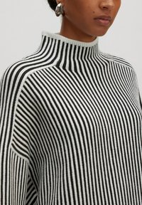 Pull à col roulé noir et blanc à rayures avec une texture côtelée, un col haut et des manches larges. Présenté avec des boucles d'oreilles métalliques.