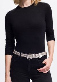 Kobieta w czarnym longsleeve'ie i czarnych spodniach z srebrnym plecionym paskiem, z jedną ręką w kieszeni na białym tle.