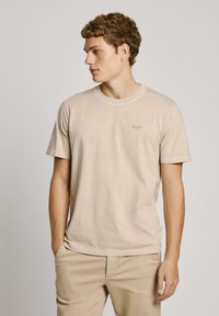 Camiseta de algodón color beige claro con cuello redondo, manga corta y un pequeño logo en el pecho izquierdo. Textura suave, ajuste casual.