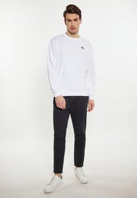 Hanorac alb cu logo, pantaloni negri slim-fit și adidași albi cu accente verzi. Modelul stă pe un fundal neutr.