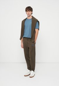 Olivgrüne Jogginghose, blaues T-Shirt und ein brauner Pullover, lässig über die Schultern gelegt. Trägt weiße Sneaker mit grauen Akzenten. Einfaches, lässiges Outfit.