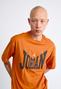 Orange bomulds t-shirt med et stort sort "JORDAN" print og en hvid logo accent, med en afslappet pasform og rund halsudskæring.