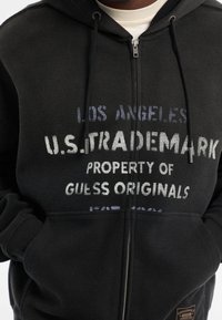Schwarze zip-up Kapuzenjacke mit strukturiertem Muster. Verfügt über einen Drucktext in verschiedenen Farbtönen und ein Logo-Patch auf der Vorderseite der Tasche. Mit Kordelzug-Kapuze.