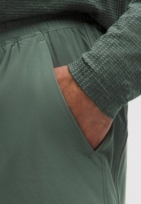 Hand in der Seitentasche von grünen dehnbaren Baumwollhosen, mit einem langärmligen, strukturierten grünen Hemd.