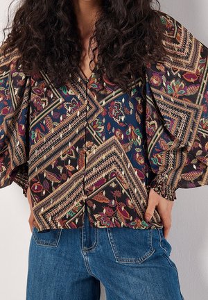 Vrouw draagt een blouse met lange mouwen met een meerkleurig bloem- en geometrisch patroon, gecombineerd met blauwe spijkerbroeken.