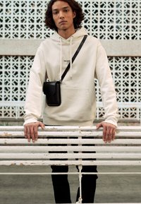 Hanorac de culoare crem cu buzunar frontal, purtat cu pantaloni negri. O geantă mică neagră de tip crossbody atârnă de o curea neagră. Fundal texturat.