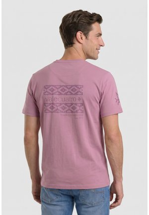 Hombre de espaldas que lleva una camiseta rosa empolvado con un logo estampado y el texto "valecuatro Tradición Argentina Est. 1971" y vaqueros azules.