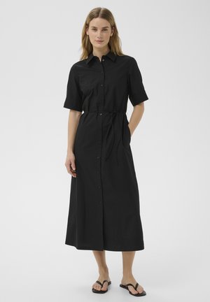 Mujer de cabello claro que lleva un vestido midi negro con botones, mangas cortas y cinturón anudado, combinado con sandalias planas negras.